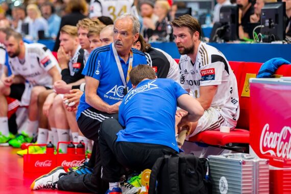 Ein Handballspieler sitzt auf einer Bank, während ein Arzt sein Bein untersucht. Andere Mannschaftsmitglieder und Mitarbeiter sitzen in der Nähe, alle tragen Uniformen. Im Hintergrund sind Sportgeräte und ein Coca-Cola-Schild zu sehen.