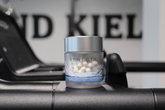 Ein kleines gefrostetes Glas mit der Aufschrift BENECTA und weißen Kapseln sitzt auf dem schwarzen Griff eines Fitnessgeräts. Der Hintergrund ist unscharf und zeigt einen großen, teilweise sichtbaren Text.