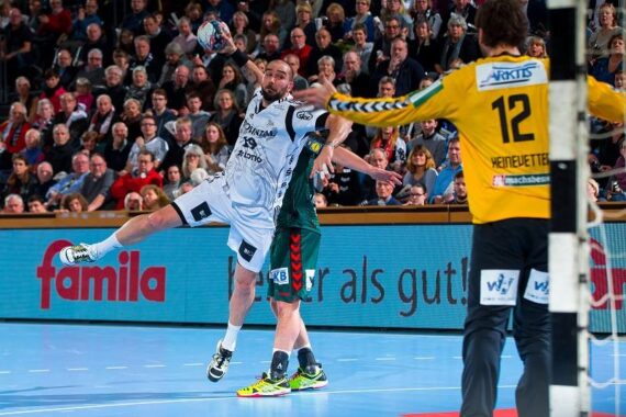 Ein Handballspieler in einem weißen Trikot springt zum Schuss, während er von einem Spieler in Grün verteidigt wird. Ein Torwart in einem gelben Trikot steht in der Nähe des Tores bereit, während im Hintergrund eine Menschenmenge zusieht.