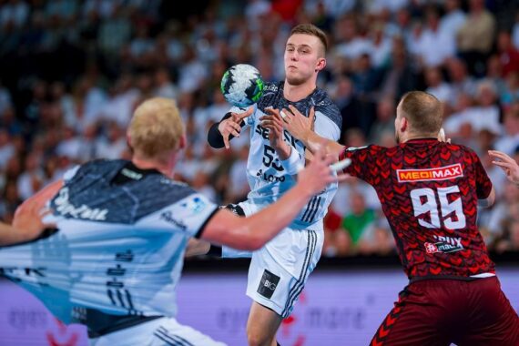 Drei männliche Handballspieler liefern sich während eines Spiels einen intensiven Wettkampf, wobei einer von ihnen im Begriff ist, einen Ball in der Luft zu fangen. Zwei Spieler tragen weiße Trikots, während der dritte, der ihnen gegenübersteht, ein rot gemustertes Trikot trägt. Im Hintergrund sieht eine unscharfe Menschenmenge zu.