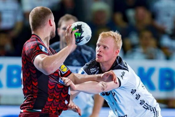 Zwei männliche Handballspieler kämpfen während eines Spiels um den Ball; der eine in einem roten Trikot hält den Ball, während der andere in einem weißen Trikot versucht, ihn zu blockieren. Im Hintergrund ist eine unscharfe Menschenmenge zu sehen.