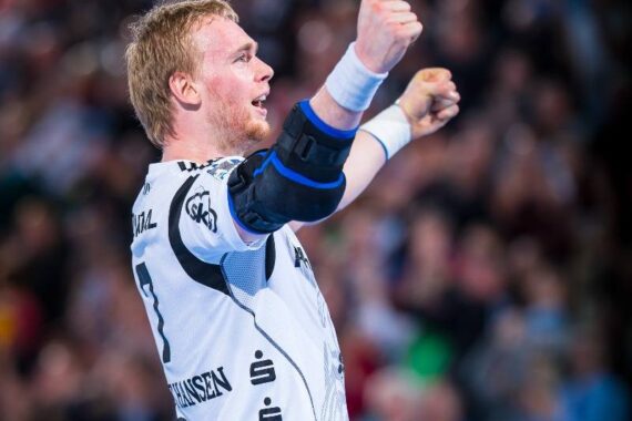 Ein männlicher Handballspieler in einem weißen Trikot reckt während eines Spiels beide Fäuste in die Höhe und jubelt.