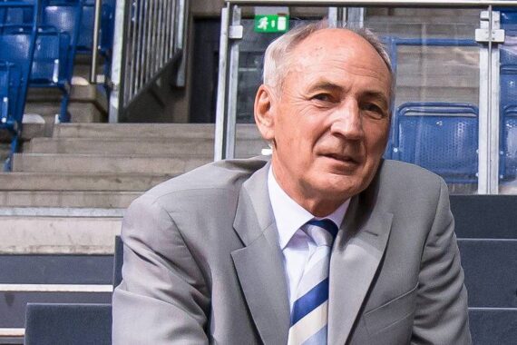 Ein älterer Mann in einem hellgrauen Anzug mit gestreifter Krawatte sitzt in einer Arena mit blauen Sitzen und Betonstufen im Hintergrund.
