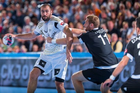 Ein Handballspieler im weißen Trikot bereitet sich darauf vor, den Ball zu werfen, während er von einem Spieler im schwarzen Trikot während eines Spiels verteidigt wird.