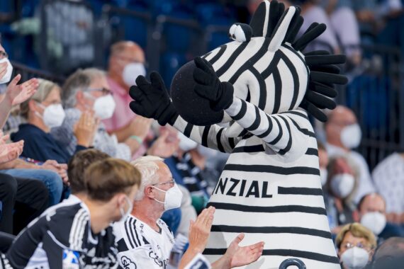 Eine Person in einem Zebra-Maskottchen-Kostüm mit der Aufschrift ANZIAL steht inmitten einer Menschenmenge, die Masken trägt, und klatscht bei einer Indoor-Veranstaltung.