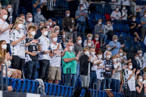 Eine große Menschenmenge mit Gesichtsmasken steht und applaudiert in einer Halle. Die meisten von ihnen tragen passende Sporttrikots, was darauf hindeutet, dass es sich um Fans einer Sportveranstaltung handelt. Blaue Sitze füllen den Hintergrund.
