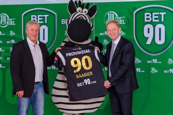 Drei Männer, einer im Zebrakostüm, posieren vor einer grünen BET 90-Kulisse. Das Maskottchen hält ein schwarzes Trikot mit der Aufschrift Provinzial 90 Saager in der Hand. Die beiden Männer auf jeder Seite lächeln.