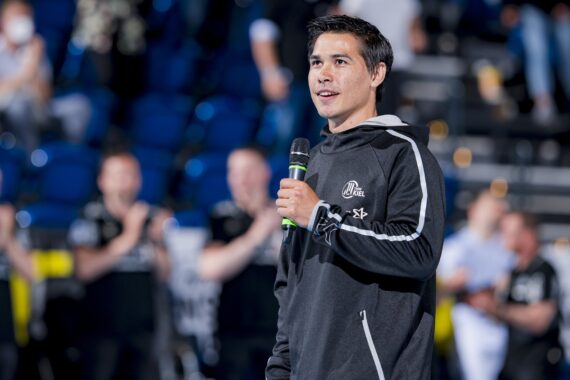 Ein Mann in einer dunklen Sportjacke hält ein Mikrofon und spricht vor einem Publikum in einer großen Halle mit blauen Stadionsitzen. Der Hintergrund ist unscharf und zeigt klatschende Menschen.