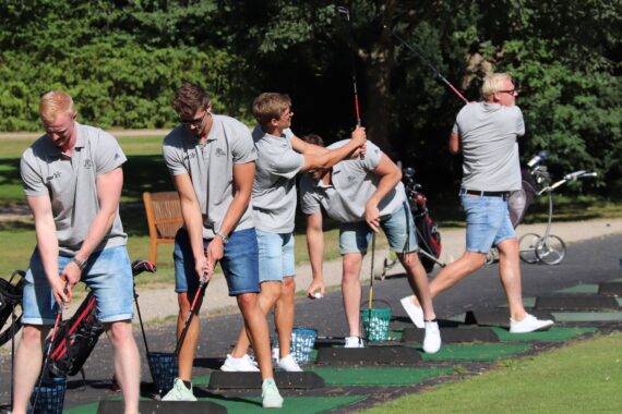 Fünf Männer in grauen Hemden und Jeans-Shorts üben an einem sonnigen Tag auf einer Driving Range ihren Golfschwung, jeder in einem anderen Stadium des Schwungs, mit Bäumen und Golfausrüstung im Hintergrund.