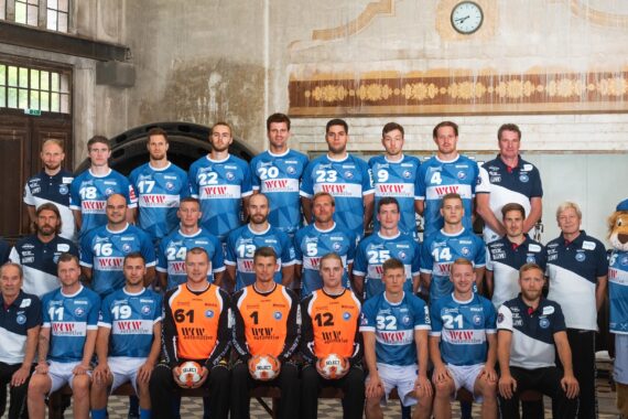 Eine Handballmannschaft in blau-weißen Trikots stellt sich in einer Halle für ein Gruppenfoto auf. Trainer und Betreuer stehen an den Seiten, und ein als Löwe gekleidetes Maskottchen steht auf der rechten Seite. Der Hintergrund hat einen alten, historischen Look.
