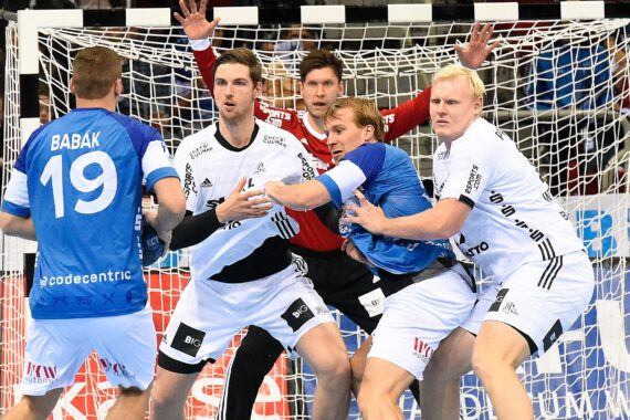 Vier Handballspieler, zwei in Blau und zwei in Weiß, stehen dicht gedrängt vor dem Tor, mit einem Torwart in Rot dahinter, der bereit ist, den Schuss zu parieren. Alle sind konzentriert und in ein intensives Spiel vertieft.