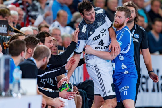 Ein Handballspieler in einem weißen Trikot wird von einem Spieler in einem blauen Trikot während eines spannenden Spiels in der Nähe einer überfüllten Bank ins Aus geschubst. Die Bankspieler der weißen Mannschaft reagieren auf diese Aktion.
