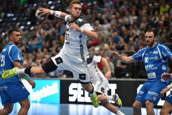 Ein Handballspieler in einem weißen Trikot springt in der Luft und bereitet sich auf einen Schuss vor, während drei Spieler in Blau versuchen, ihn während eines Spiels in einer überfüllten Halle zu blockieren.