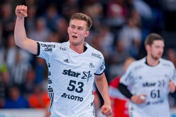 Ein männlicher Handballspieler in einem weißen Trikot mit der Nummer 53 hebt jubelnd die rechte Faust auf dem Spielfeld, während im Hintergrund ein anderer Spieler und eine Menschenmenge zu sehen sind.