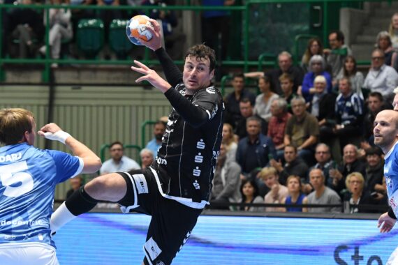 Ein Handballspieler in einem schwarzen Trikot springt, um den Ball zu schießen, umgeben von gegnerischen Spielern in blauen Trikots, während eine Menschenmenge das Spiel in einer Halle verfolgt.