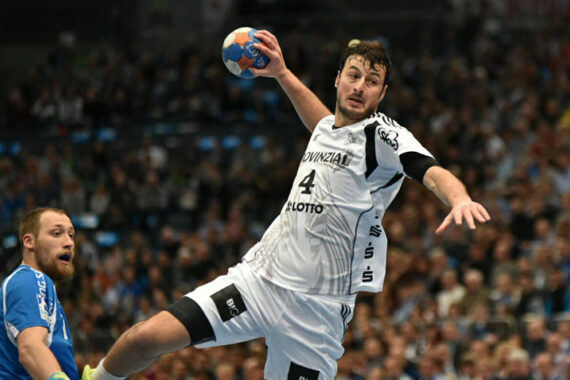 Ein männlicher Handballspieler in einem weißen Trikot springt in der Luft, um den Ball zu werfen. Ein Verteidiger in einer blauen Uniform steht neben ihm. Das Geschehen spielt sich in einer Halle mit einer unscharfen Menschenmenge im Hintergrund ab.