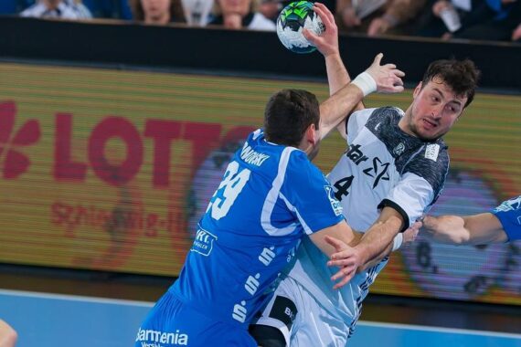 Ein Handballspieler in einem weiß-schwarzen Trikot versucht einen Schuss, während er von einem Spieler in einem blauen Trikot während eines Spiels eng verteidigt wird. Der Hintergrund zeigt eine hell erleuchtete Arena und Zuschauer.