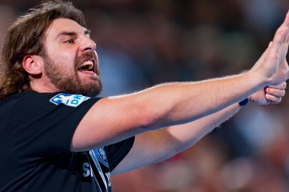Ein Mann mit langen Haaren und Bart gestikuliert während einer Sportveranstaltung leidenschaftlich mit ausgestreckten Armen und offenem Mund, als würde er Anweisungen oder Ermutigungen geben. Er trägt ein dunkles Hemd.