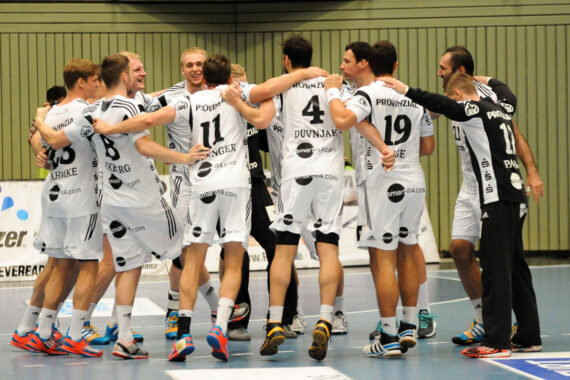 Eine Gruppe von männlichen Handballspielern in weißen Trikots steht im Kreis, die Arme umeinander gelegt, und feiert auf einem Hallenplatz. Sie lächeln und wirken fröhlich, einige springen im Gleichschritt.