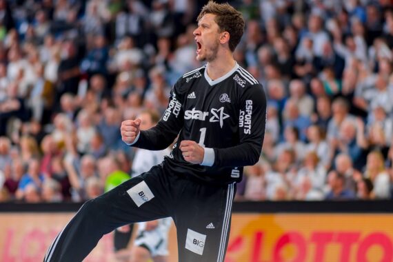 Ein männlicher Handballspieler in einem schwarzen Trikot, der mit geballten Fäusten und offenem Mund vor einer jubelnden Menge in einer Sporthalle energisch jubelt.