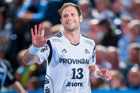 Ein männlicher Handballspieler in einem weißen Trikot mit der Nummer 13 lächelt und winkt mit seiner rechten Hand während eines Spiels. Im Hintergrund ist eine unscharfe Menschenmenge zu sehen.