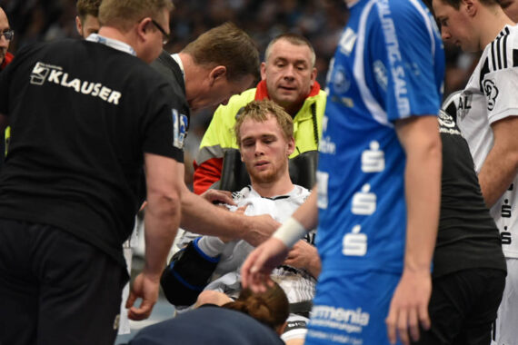 Ein männlicher Sportler in einem weißen Trikot sitzt auf dem Boden und scheint verletzt zu sein, während Sanitäter und andere Personen ihn umringen, um ihm zu helfen. Menschen in schwarzen und blauen Uniformen stehen in der Nähe und zeigen sich besorgt.