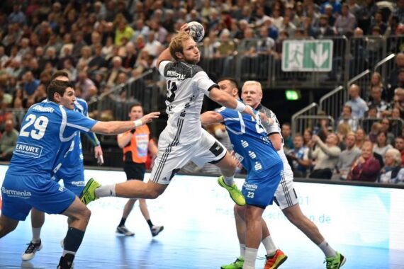 Ein Handballspieler in einem weißen Trikot springt, um den Ball zu werfen, während drei Verteidiger in Blau versuchen, ihn während eines Hallenspiels zu blockieren, während im Hintergrund eine große Menschenmenge zusieht.