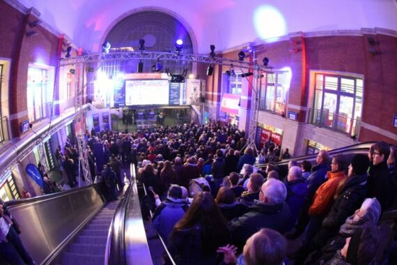 Eine große Menschenmenge versammelt sich in einer hell erleuchteten Halle, vor einer Bühne mit großen Bildschirmen und Beleuchtungsanlagen. Die Menschen stehen in der Nähe einer Rolltreppe und auf den Balkonen und beobachten das Geschehen unten.