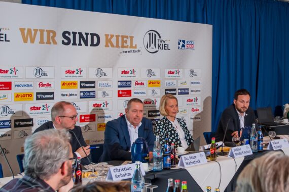 Vier Personen sitzen an einem Konferenztisch mit Mikrofonen und Namensschildern vor einer Kulisse mit dem Logo des THW Kiel und Sponsorenlogos. Darüber prangt ein Banner mit der Aufschrift "WIR SIND KIEL". Im Vordergrund sind Fotografen und Anwesende zu sehen.
