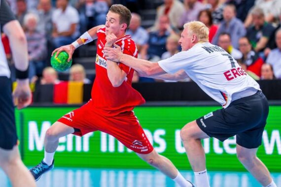 Ein Handballspieler in einem roten Trikot versucht, einen Verteidiger in einem weißen Trikot, der ihn blockieren will, während eines intensiven Spiels zu überholen.