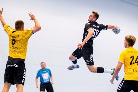 Ein männlicher Handballspieler in einem schwarzen Trikot springt in der Luft, um den Ball zu werfen, während zwei gegnerische Spieler in gelben Trikots versuchen, ihn zu blockieren. Im Hintergrund ist ein Offizieller in Blau zu sehen.