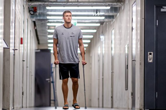 Ein junger Mann in einem grauen T-Shirt, schwarzen Shorts und Sandalen geht auf Krücken durch einen hell erleuchteten Flur mit weißen Wänden und freiliegenden Rohren über ihm.