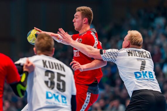 Drei männliche Handballspieler treten während eines Spiels gegeneinander an. Ein Spieler in Rot springt mit dem Ball, während zwei Spieler in weißen Trikots, die mit Kuhn (35) und Wiencek (7) bezeichnet sind, versuchen, ihn zu blockieren. Im Hintergrund ist ein unscharfes Publikum zu sehen.