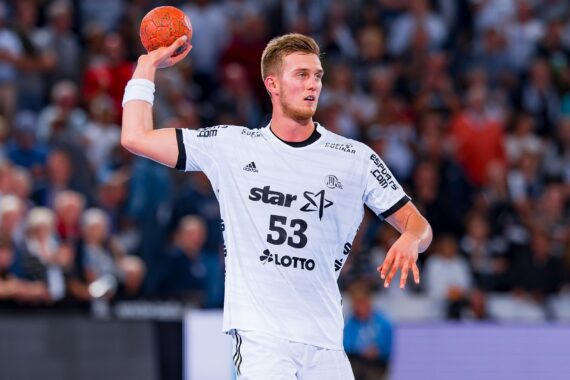Ein Handballspieler, der ein weißes Trikot mit der Nummer 53 trägt, hält während eines Spiels in einer überfüllten Halle einen orangefarbenen Ball über seinen Kopf und bereitet sich auf den Wurf vor.