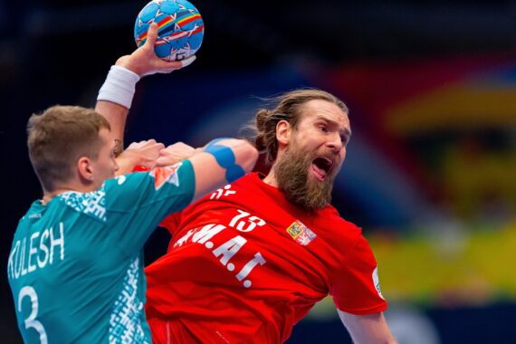 Ein männlicher Handballspieler in einem roten Trikot springt und wirft den Ball, während ein Verteidiger in einem blaugrünen Trikot versucht, ihn während eines intensiven Spiels zu blockieren.