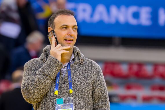 Ein Mann, der einen grauen Strickpullover und ein Veranstaltungsabzeichen trägt, spricht in einer Halle mit blauen und roten Sitzgelegenheiten im Hintergrund auf sein Smartphone.