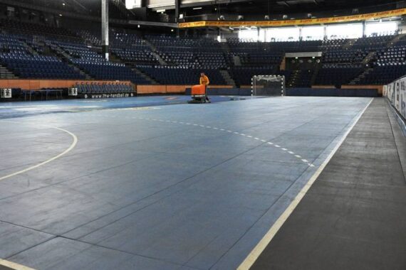 Eine Person bedient eine Bodenreinigungsmaschine in einer leeren Sporthalle mit blauem Bodenbelag, umgeben von Reihen blauer Stadionsitze.