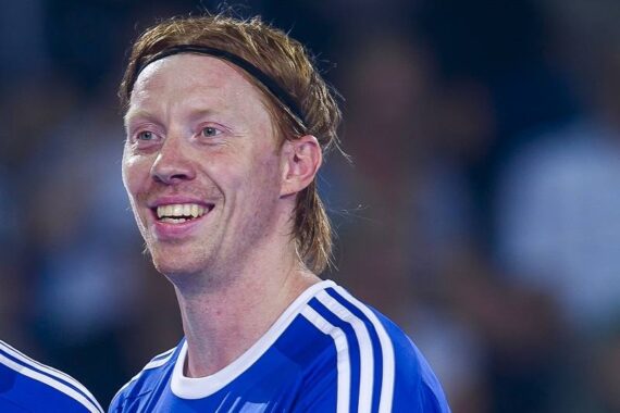 Ein lächelnder Sportler mit heller Haut, rotem Haar und einem schwarzen Stirnband, der ein blau-weißes Sporttrikot trägt, steht in einer Halle.