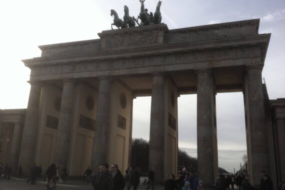 Das Bild zeigt das Brandenburger Tor in Berlin, Deutschland, mit Menschen, die an einem bewölkten Tag vor dem historischen Monument spazieren gehen und sich versammeln.