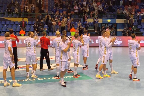 Eine Männer-Handballmannschaft in weißen Trikots steht auf einem Spielfeld und klatscht dem Publikum zu. Einige Spieler sind den Zuschauern zugewandt, andere sind abgewandt, während im Hintergrund Fans zu sehen sind, die von der Tribüne aus jubeln.