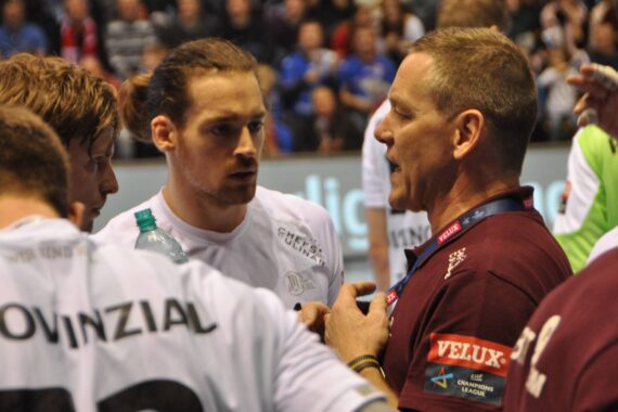 Ein Trainer in einem kastanienbraunen Hemd spricht während einer Pause bei einem Handballspiel zu drei männlichen Sportlern in weißen Trikots. Die Spieler hören aufmerksam zu, einer von ihnen hält eine Wasserflasche in der Hand. Im Hintergrund ist eine unscharfe Menschenmenge zu sehen.