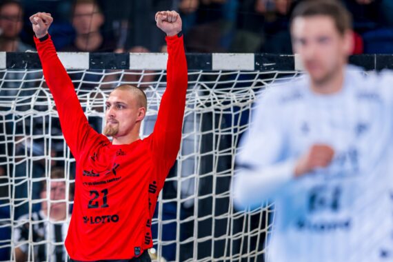 Ein Handballtorwart in einem roten Trikot steht mit feierlich erhobenen Armen vor einem Tornetz, während im Vordergrund ein unscharfer Spieler in einem weißen Trikot zu sehen ist.