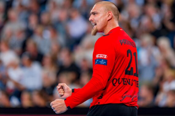 Ein männlicher Handballspieler in einem roten Trikot ballt während eines Spiels die Fäuste und jubelt, während im Hintergrund eine unscharfe Menschenmenge zu sehen ist.