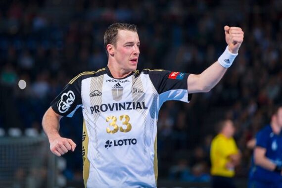 Ein männlicher Handballspieler in einem weiß-schwarzen Trikot mit der Nummer 33 jubelt mit geballter Faust während eines Spiels. Im Hintergrund sind unscharfe Zuschauer und andere Spieler zu sehen.