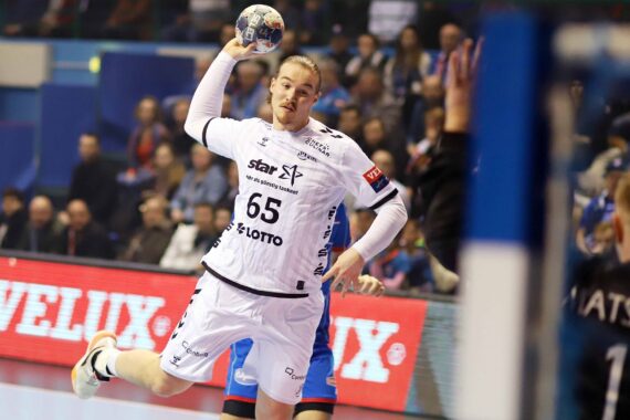 Ein Handballspieler in einem weißen Trikot bereitet sich mit erhobenem Arm und dem Ball in der Hand auf einen Torschuss während eines Hallenspiels vor. Im Hintergrund sind Gegner und Zuschauer zu sehen.