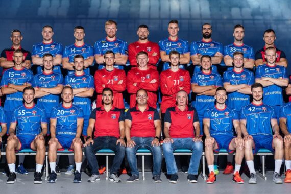 Eine Handballmannschaft der Männer stellt sich in einer Halle für ein Gruppenfoto auf. Die Spieler tragen blaue Trikots, während Trainer und Betreuer in roten Hemden in vier Reihen zwischen ihnen sitzen und stehen. Alle blicken nach vorne und lächeln.