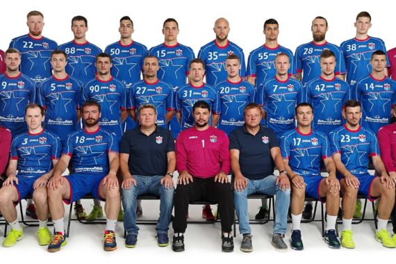 Eine Herrenhandballmannschaft stellt sich für ein Gruppenfoto auf. Die Spieler tragen blaue Trikots mit roten und weißen Akzenten, während Trainer und Mitarbeiter in Freizeitkleidung in der ersten Reihe sitzen. Alle stehen mit dem Gesicht nach vorne vor einem weißen Hintergrund.