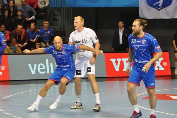 Zwei Handballspieler in blauen Trikots verteidigen sich während eines Spiels gegen einen Spieler in einem weißen Trikot, während die Zuschauer von der Tribüne im Hintergrund zusehen. Der Boden ist für das Handballfeld markiert.