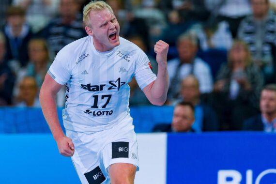 Ein männlicher Handballspieler in einem weißen Trikot mit der Nummer 17 ballt die Faust und schreit vor Freude. Er befindet sich auf einem Hallenplatz, mit unscharfen Zuschauern im Hintergrund.