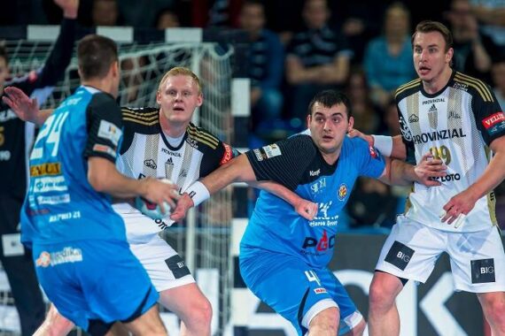 Vier männliche Handballspieler, zwei in blauen und zwei in weißen Trikots, kämpfen intensiv in der Nähe des Tores, während ein Torwart in Schwarz aus dem Hintergrund zusieht. Die Spieler wirken während des Spiels konzentriert.